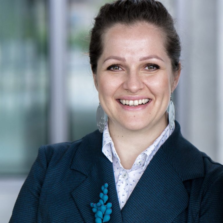 Nastya schaut strahlend in die Kamera. Sie trägt ihr dunkelblondes Haar zurückgesteckt. Nastya trägt einen dunkelblauen, glänzenden Blazer und einen hellblauen Perlen-Anstecker auf der linken Krämpe. Darunter ist ein weiß-gepunktetes Hemd zu sehen.