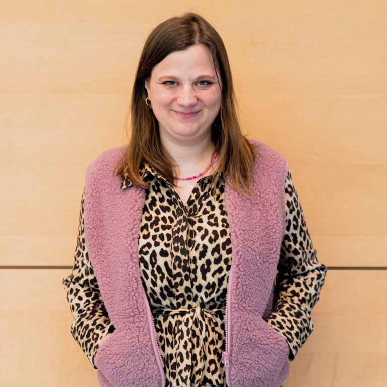Hannah Harding trägt eine Bluse mit Leopardenmuster und eine rosa Weste in Schafffelloptik sowie eine rosa-orange Kette. Hannah hat halblange goldbraune Haare und lächelt in die Kamera. Hannah steht vor einer holzfarbenen Wand.