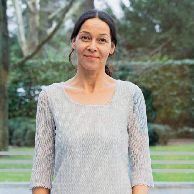 Bild Judith korrigiert Judith Gardner steht vor einer Fensterfront vor grünem Hintergrund mit Bäumen. Sie trägt einen hellgrauen, gestrickten Pullover mit weitem Halsausschnitt und dreiviertel Ärmeln. Judith hat dunkles Haar, das nach hinten gebunden ist, und lächelt leicht in die Kamera. Der Hintergrund zeigt eine natürliche Parklandschaft oder einen Garten mit Bäumen und etwas Rasen im unteren Bildbereich.