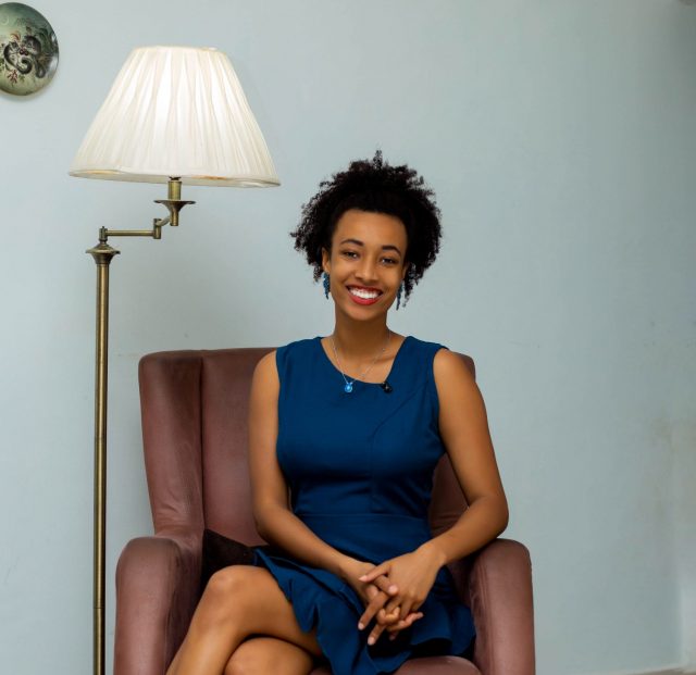 Bild Phini Phinissia sitzt in einem rot-braunen Ledersessel neben einer Stehlampe. Sie trägt ein dunkelblaues Kleid, hat die Hände entspannt auf dem Schoß gefaltet und lächelt herzlich in die Kamera.