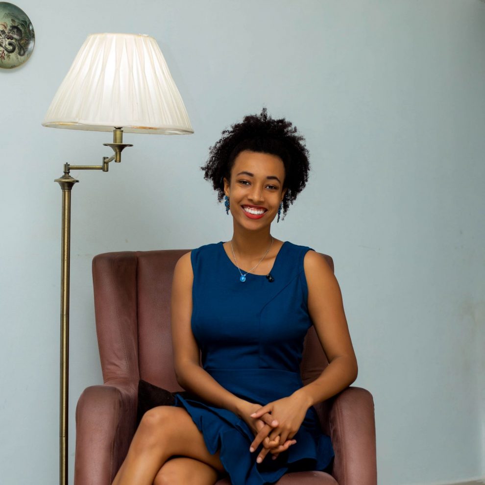 Phinissia sitzt in einem rot-braunen Ledersessel neben einer Stehlampe. Sie trägt ein dunkelblaues Kleid, hat die Hände entspannt auf dem Schoß gefaltet und lächelt herzlich in die Kamera.