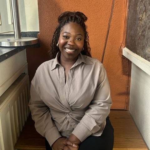 Tracy sitzt in einer Nische vor einem organgenen Hintergrund. Sie trägt eine hellbraune, lange Bluse und goldene Ohrringe und lächelt in die Kamera.