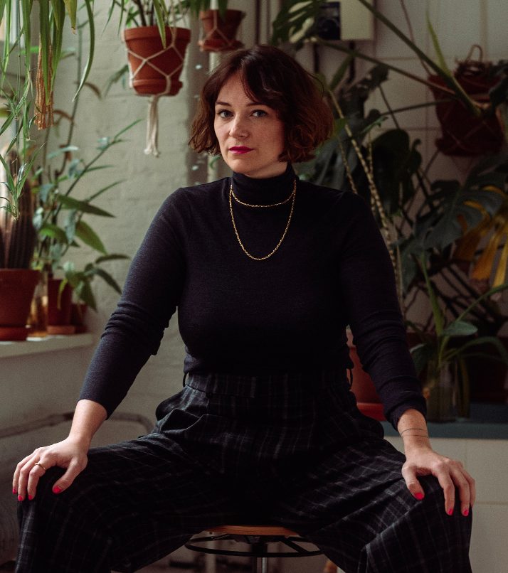 Mareike Sitzt in schwarzem Langarmshirt und schwarzer langer Hose auf einem Hocker. Die Hände hat sie auf den Knien abgestützt. Mareice schaut im Profil in die Kamera. Die dunklen Haare trägt sie zu einem offenen Bob, der auf Kinnhöhe endet. Im Hintergrund sind Pflanzen zu sehen.