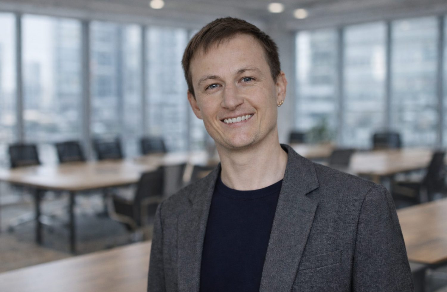 né schaut lächelnd in die Kamera. Né trägt ein graues Sakko und steht in einem Büro.