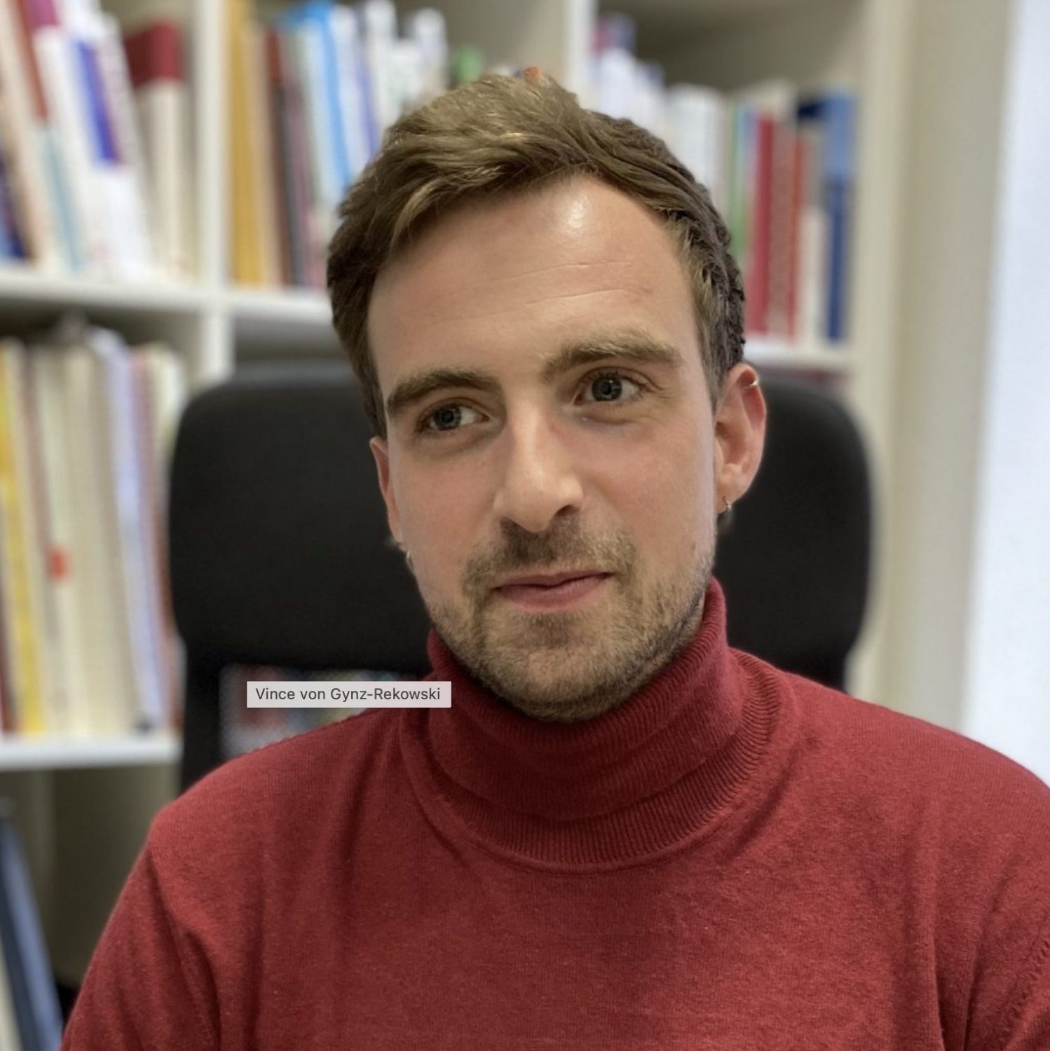 Vince trägt einen dunkelroten Rollkragenpullover. Er sitzt vor einem bunten Bücherregal. Vince schaut leicht links neben die Foto-Linse und schaut neutral freundlich.