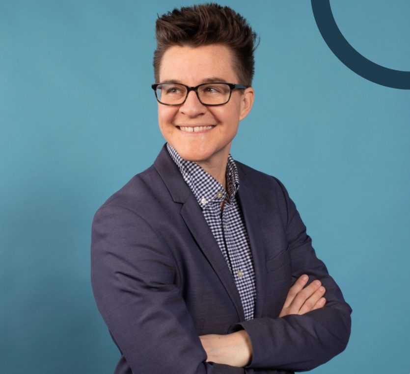 Erica steht vor einer blauen Wand. Erica trägt einen blauen Blazer, ein kariertes Hemd und eine dunkelumrahmte Brille. Ihre Haare trägt sie kurz.
