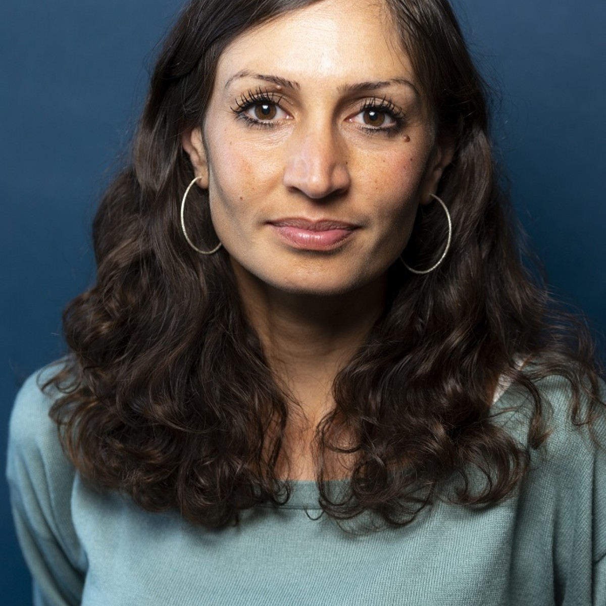 Gilda Sahebi schaut direkt in die Kamera. Sie trägt ein türkisfarbenes langarm-Oberteil. Ihre langen, dunkelbraunen Locken trägt sie offen. Ihre Ohren schmücken große Kreolen.