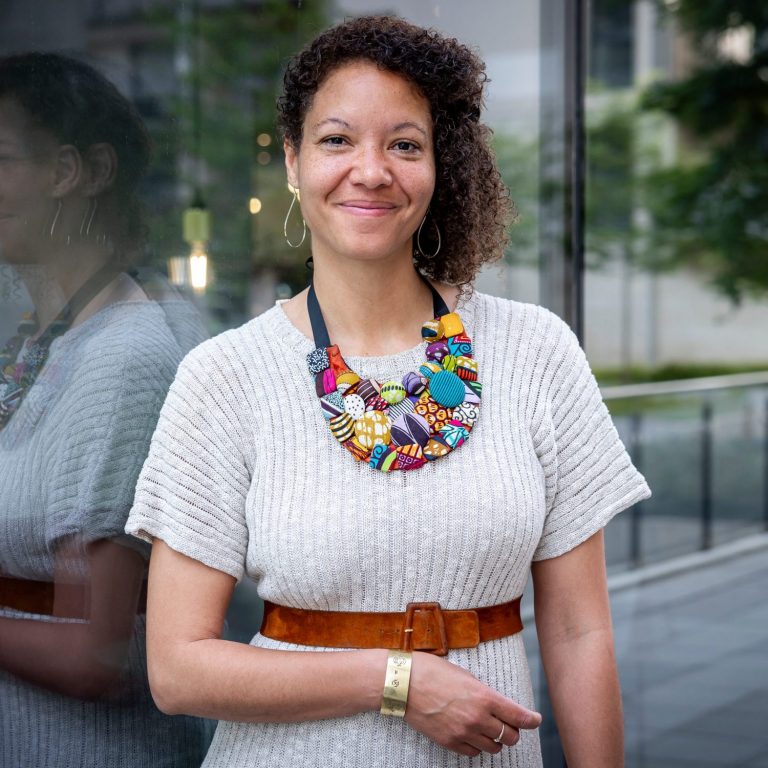 Dr. Hedda Ofoole Knoll lächelt freundlich in die Kamera. Hedda trägt ein hellgraues, kurzärmeliges Strickkleid mit einem braunen Ledergürtel und eine auffällige, bunte Halskette aus verschiedenen gemusterten Stoffelementen. Heddas lockige Haare fallen über die Schultern und Hedda trägt große goldene Kreolen-Ohrringe sowie ein goldenes Armband. Im Hintergrund sind Glasfronten und grüne Pflanzen eines modernen Gebäudes zu sehen.