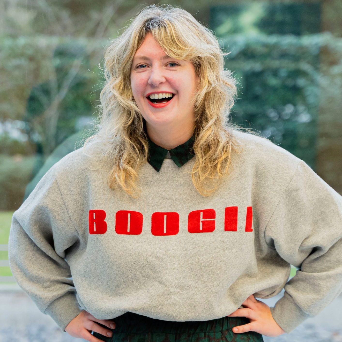 In diesem Bild ist Lucy Jo Fitz zu sehen. Lucy hat langes, welliges blondes Haar und steht lächelnd vor einer Fensterscheibe mit Blick ins Grüne. Lucy trägt einen grauen Pullover mit dem roten Schriftzug "BOOGI!" auf der Brust. Darunter ist ein dunkles Oberteil mit Kragen erkennbar. Im Hintergrund sind verschwommen grüne Pflanzen oder Bäume zu sehen. Lucy hat die Hände in die Hüften gestemmt und lacht herzlich in die Kamera.