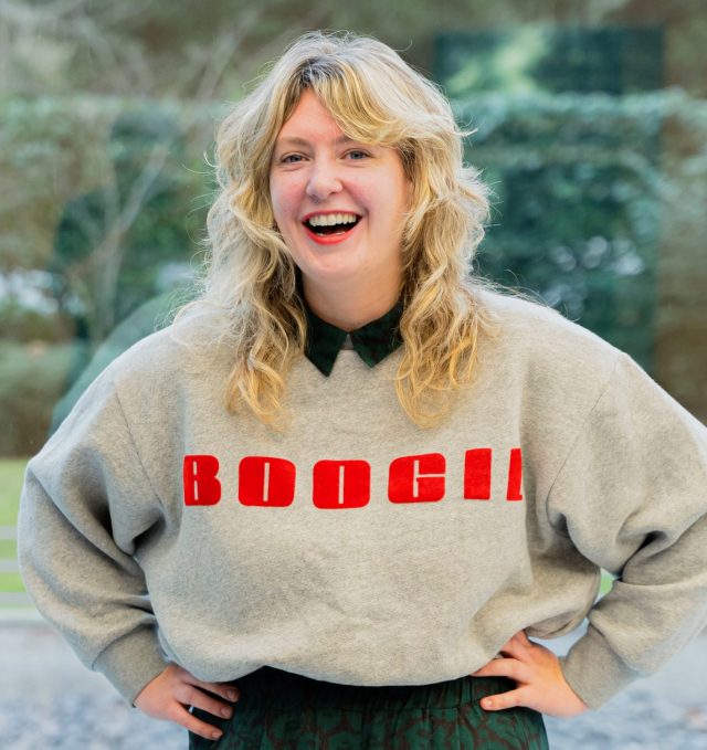 In diesem Bild ist Lucy Jo Fitz zu sehen. Lucy hat langes, welliges blondes Haar und steht lächelnd vor einer Fensterscheibe mit Blick ins Grüne. Lucy trägt einen grauen Pullover mit dem roten Schriftzug "BOOGI!" auf der Brust. Darunter ist ein dunkles Oberteil mit Kragen erkennbar. Im Hintergrund sind verschwommen grüne Pflanzen oder Bäume zu sehen. Lucy hat die Hände in die Hüften gestemmt und lacht herzlich in die Kamera.