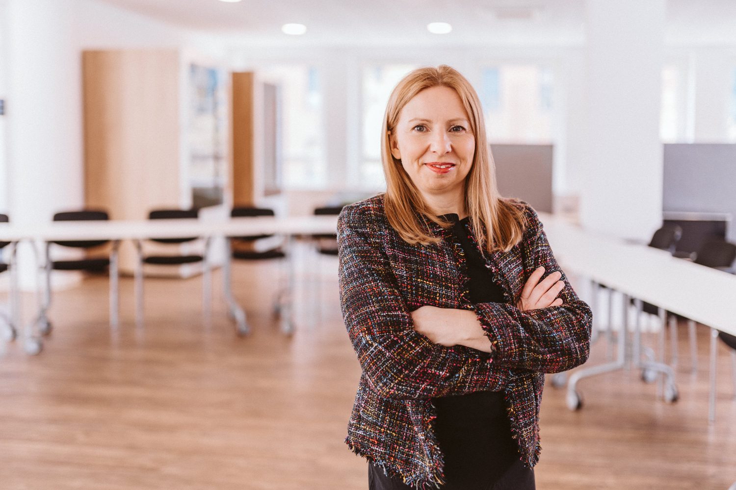 Nadine steht in einem hellen Büro. Sie trägt ihre rotblonden Haare offen und hat ihre Arme verschränkt. Sie lächelt leicht in die Kamera.