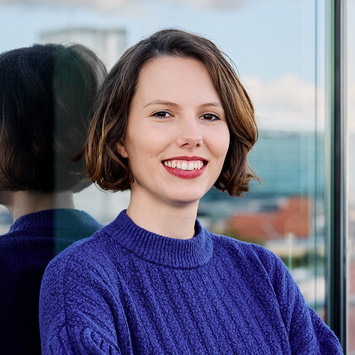 Vera lächelt zähneblitzend in die Kamera. Sie lehnt vor einer Glasscheibe, trägt ihre braunen Haare offen und auf Kinnlänge. Ihre Lippen schmückt ein dunkelroter Lippenstift und sie trägt einen dunkelblauen Wollpullover.